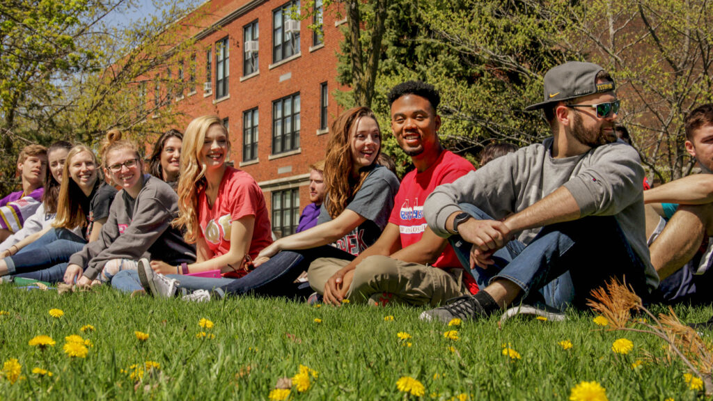 Students in Greek letter shirts sitting on a green lawn on campus